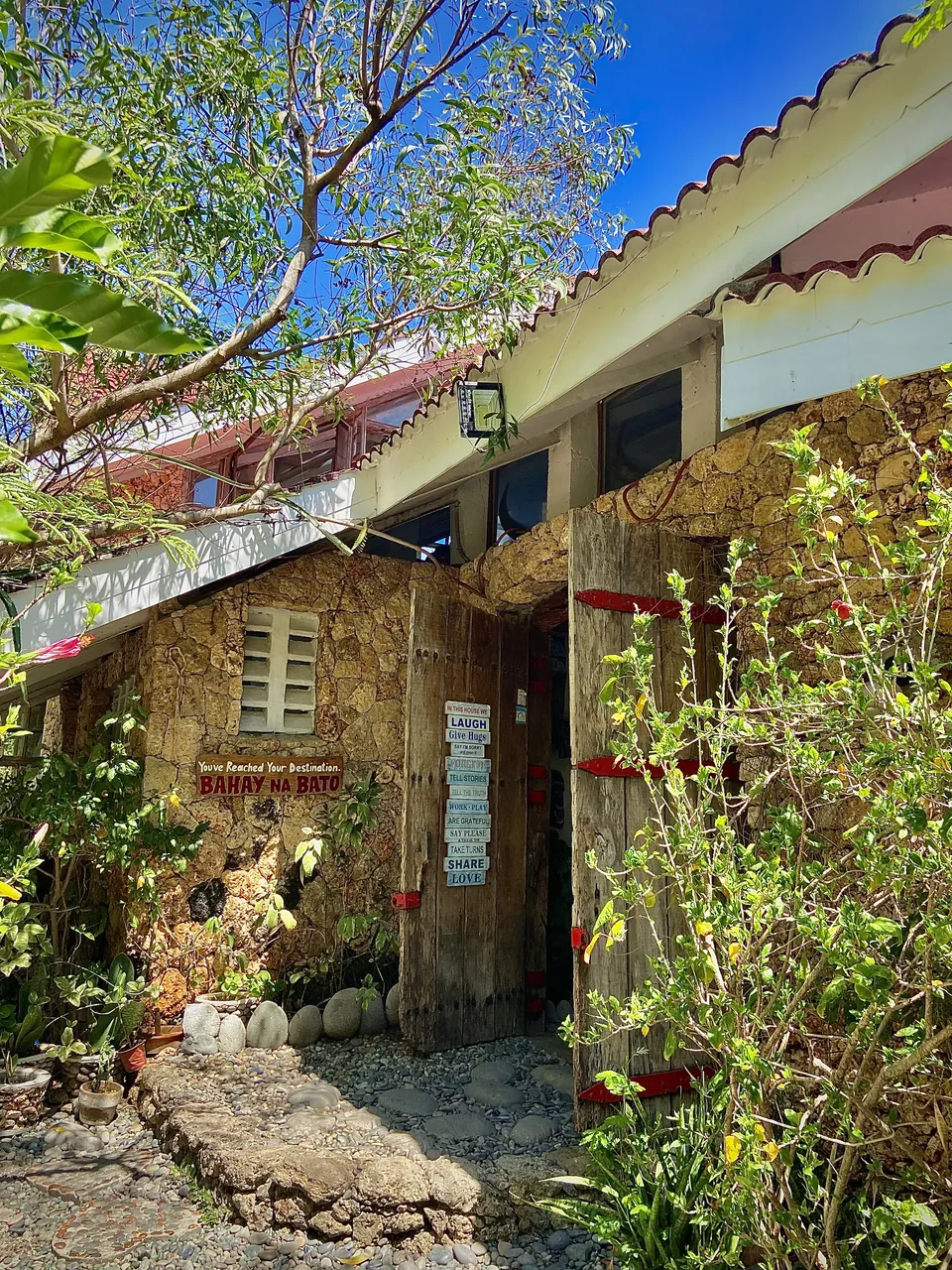 The stone exterior and open timber doorway of Bahay na Bato, framed by garden trees