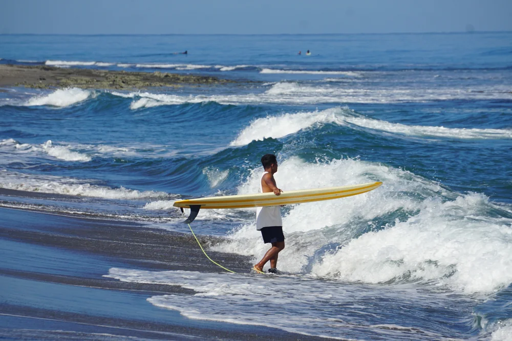 A surfer carrying a yellow longboard beside the rolling waves of Urbiztondo Beach